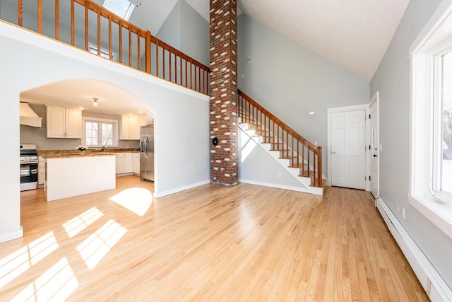 unfurnished living room with arched walkways, stairway, baseboard heating, light wood-style floors, and baseboards