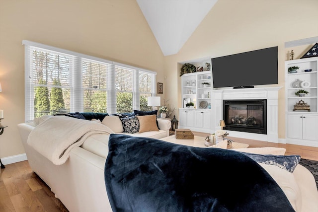living room featuring built in features, wood finished floors, baseboards, high vaulted ceiling, and a glass covered fireplace