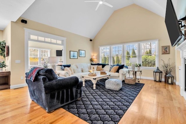 living room featuring light wood-style floors, high vaulted ceiling, a glass covered fireplace, a ceiling fan, and baseboards