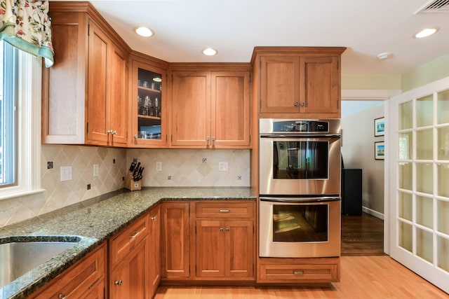 kitchen featuring stainless steel double oven, stone counters, visible vents, brown cabinets, and glass insert cabinets