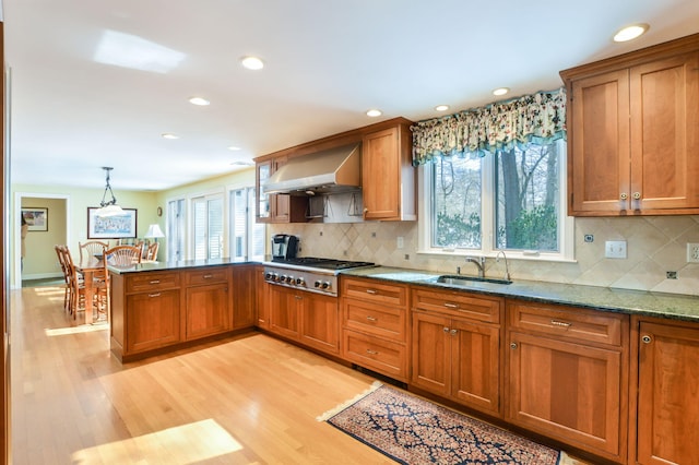 kitchen featuring light wood-style flooring, brown cabinets, wall chimney range hood, stainless steel gas cooktop, and a sink