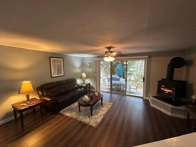 living room featuring wood finished floors, ceiling fan, baseboards, a wood stove, and a textured ceiling