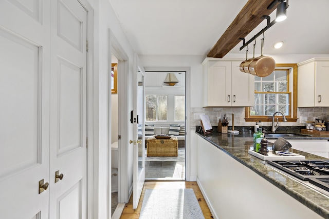 kitchen with dark stone countertops, beam ceiling, a sink, white cabinetry, and backsplash