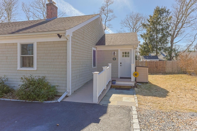 view of exterior entry featuring a chimney, roof with shingles, and fence