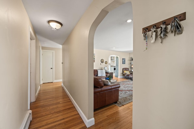 corridor with wood finished floors, arched walkways, baseboard heating, and baseboards