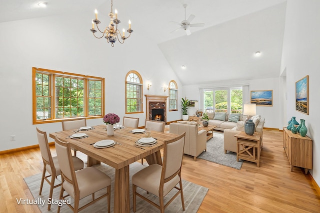 dining space featuring high vaulted ceiling, light hardwood / wood-style flooring, a brick fireplace, and ceiling fan with notable chandelier