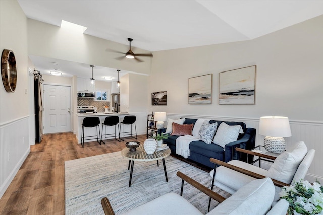 living room with vaulted ceiling, light wood-style flooring, a ceiling fan, and wainscoting