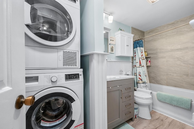 washroom featuring stacked washer / drying machine, wainscoting, wood finished floors, and laundry area