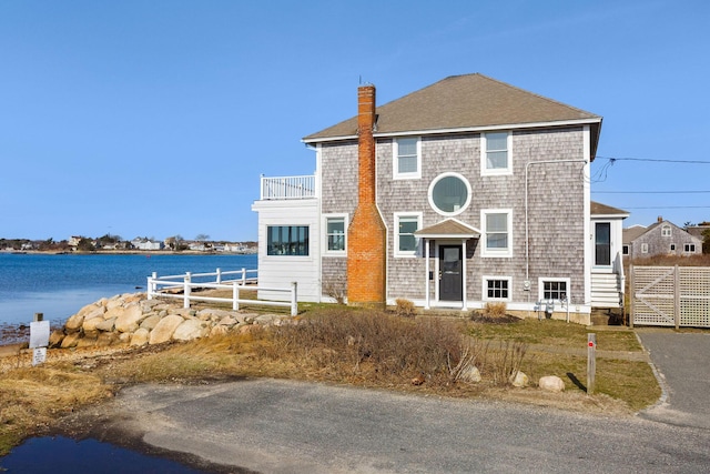 view of front of house featuring a chimney, fence, a balcony, a shingled roof, and a water view