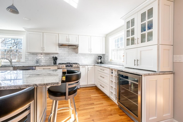 kitchen featuring beverage cooler, tasteful backsplash, light wood-style floors, a breakfast bar, and under cabinet range hood