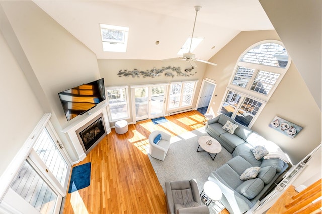 living room with a skylight, a ceiling fan, a glass covered fireplace, wood finished floors, and high vaulted ceiling