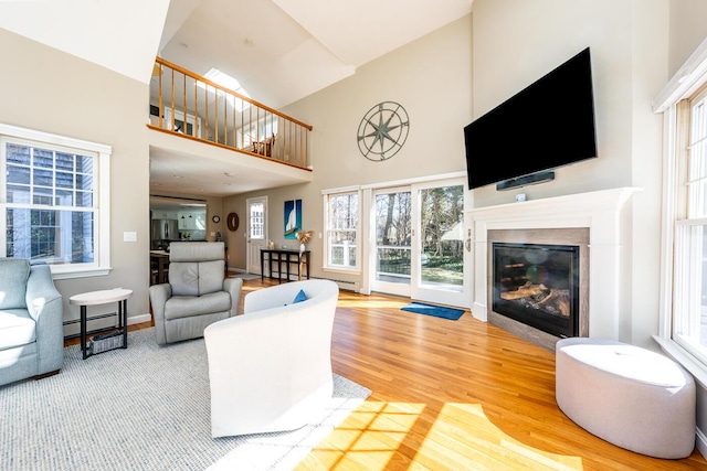 living room featuring a baseboard heating unit, wood finished floors, a towering ceiling, baseboards, and a glass covered fireplace