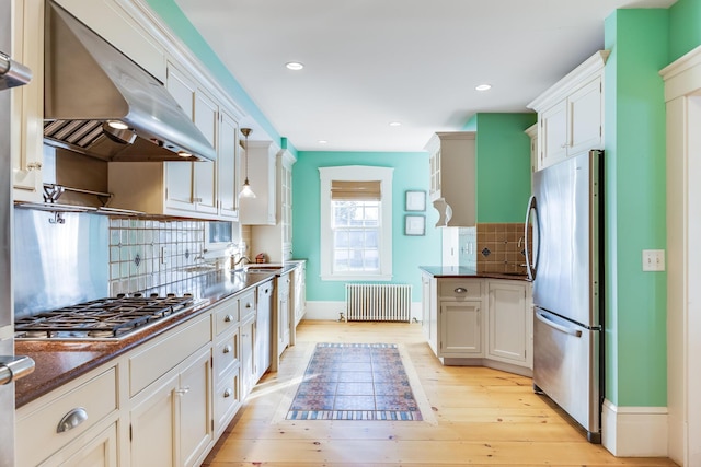 kitchen featuring white cabinetry, ventilation hood, hanging light fixtures, appliances with stainless steel finishes, and radiator heating unit