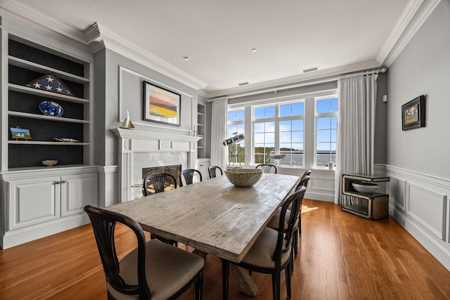 dining space with ornamental molding, built in shelves, a high end fireplace, wood finished floors, and a wainscoted wall