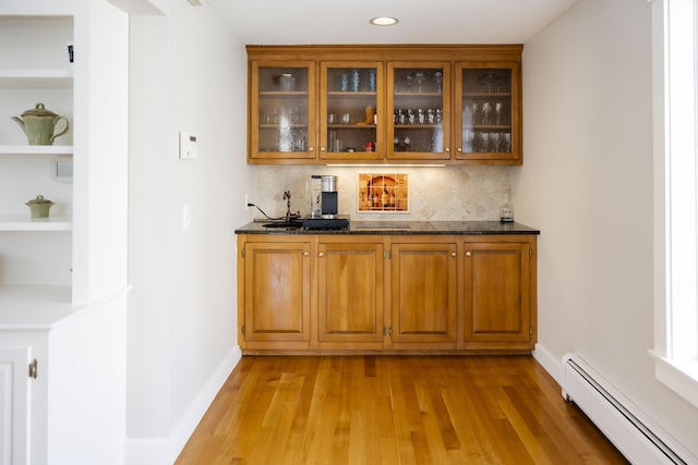 bar featuring dark stone countertops, backsplash, baseboard heating, and light hardwood / wood-style flooring