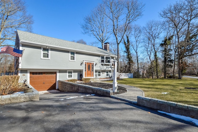 bi-level home featuring aphalt driveway, an attached garage, a shingled roof, a front lawn, and a chimney