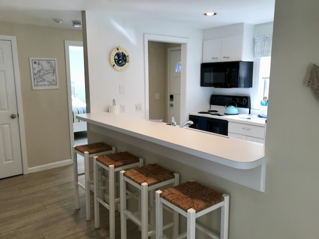 kitchen featuring black microwave, range with electric cooktop, a breakfast bar area, wood finished floors, and white cabinetry