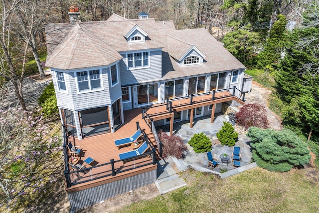 back of house with a deck, a patio, a chimney, view of wooded area, and roof with shingles
