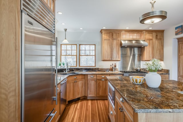 kitchen featuring built in appliances, a sink, extractor fan, light wood-style floors, and dark stone counters