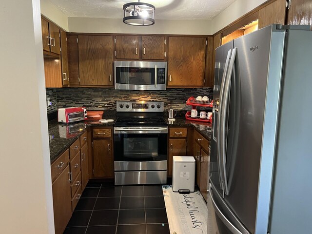 kitchen with a textured ceiling, tasteful backsplash, dark tile patterned flooring, dark stone countertops, and stainless steel appliances