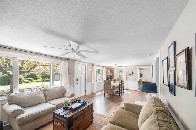 living room featuring a ceiling fan, light wood-style flooring, a textured ceiling, plenty of natural light, and a wainscoted wall