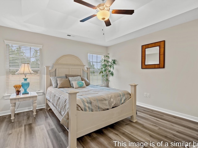 bedroom with wood-type flooring, a tray ceiling, multiple windows, and ceiling fan