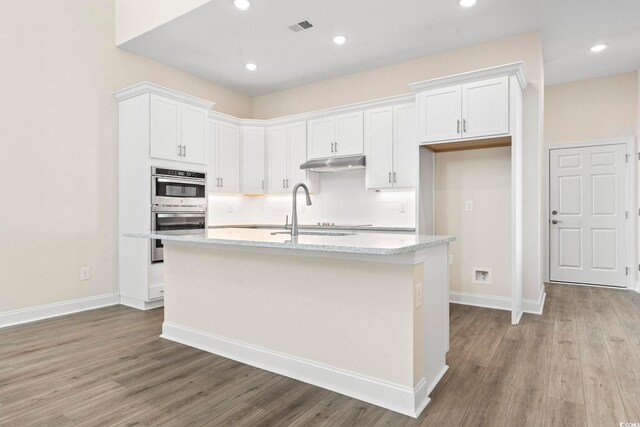 kitchen with light stone countertops, double oven, sink, a center island with sink, and white cabinetry