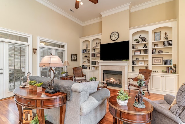 living room with built in shelves, ceiling fan, a towering ceiling, light wood-type flooring, and ornamental molding