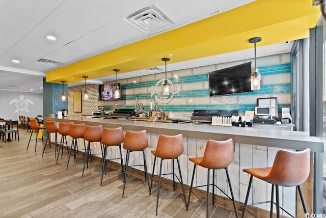 bar with a notable chandelier, a drop ceiling, stainless steel counters, hanging light fixtures, and light wood-type flooring