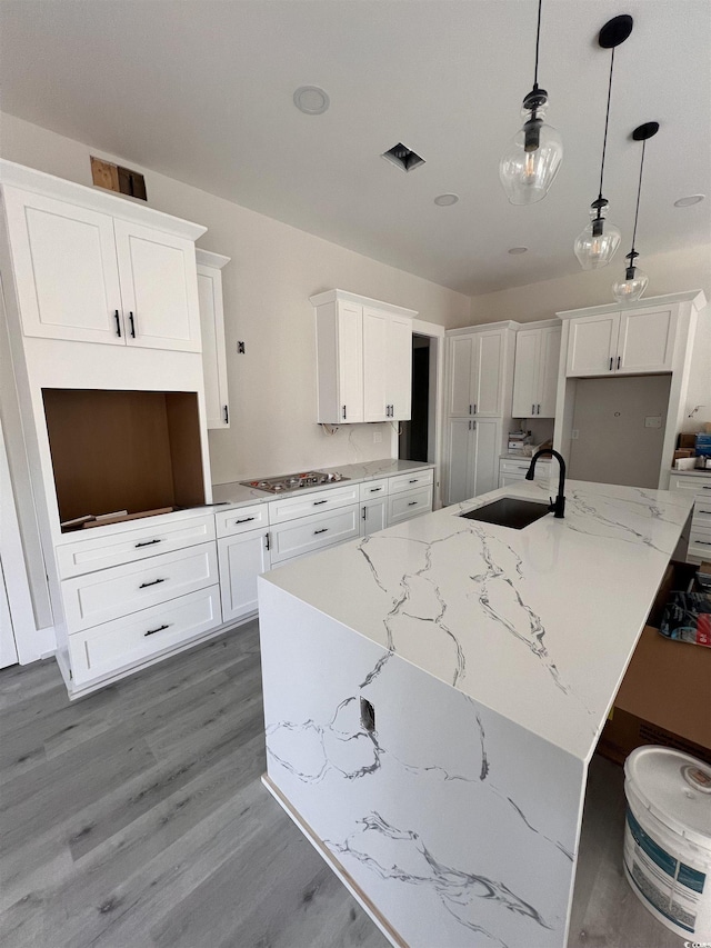 kitchen with stainless steel cooktop, wood finished floors, an island with sink, a sink, and white cabinetry