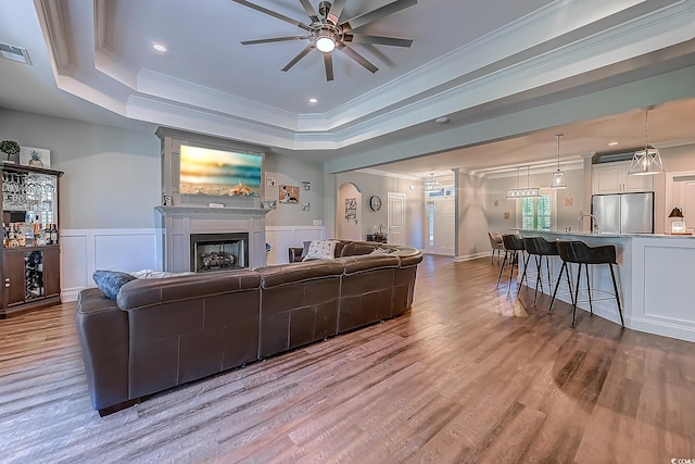 living room featuring ceiling fan, a tray ceiling, ornamental molding, light hardwood / wood-style flooring, and sink