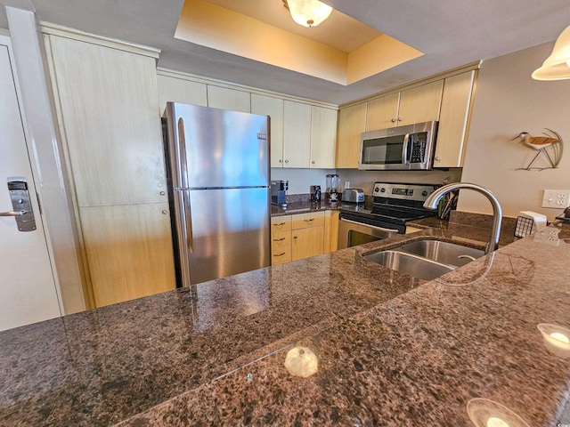 kitchen featuring light brown cabinets, a raised ceiling, appliances with stainless steel finishes, sink, and dark stone countertops