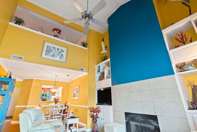 living room featuring ceiling fan, high vaulted ceiling, a tile fireplace, built in features, and crown molding