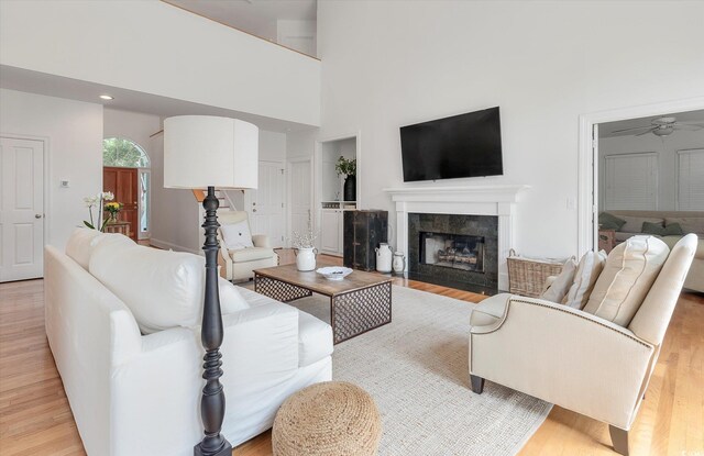 living room with ceiling fan, a tiled fireplace, light hardwood / wood-style flooring, and a towering ceiling