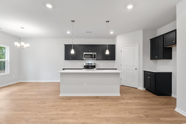 kitchen with light wood-type flooring, stainless steel appliances, hanging light fixtures, and an island with sink