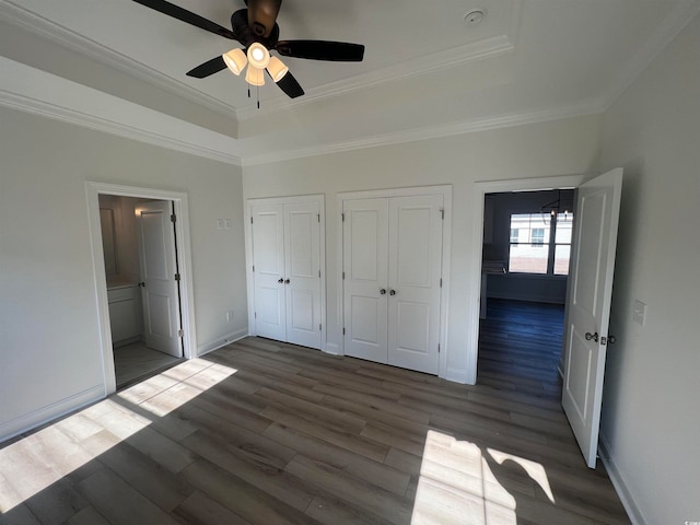 unfurnished bedroom with ornamental molding, ceiling fan, a tray ceiling, multiple closets, and dark hardwood / wood-style flooring