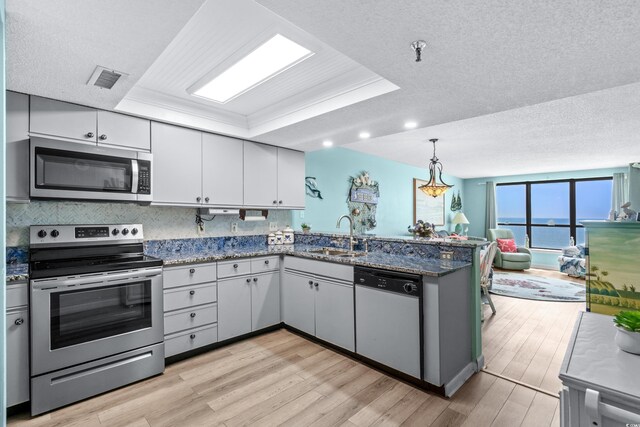 kitchen with dark stone counters, a tray ceiling, appliances with stainless steel finishes, light hardwood / wood-style floors, and kitchen peninsula