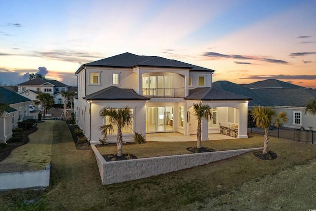 back house at dusk featuring a patio area, a lawn, and a balcony