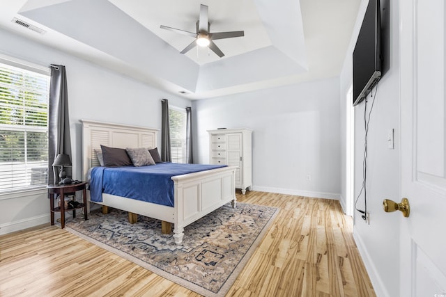 bedroom with ceiling fan, a raised ceiling, light wood-type flooring, and multiple windows