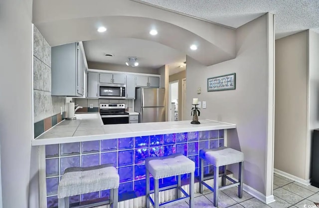 kitchen with kitchen peninsula, appliances with stainless steel finishes, gray cabinetry, light tile patterned floors, and tile countertops