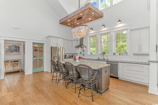 kitchen with high vaulted ceiling, stainless steel appliances, light hardwood / wood-style flooring, wall chimney range hood, and a kitchen island