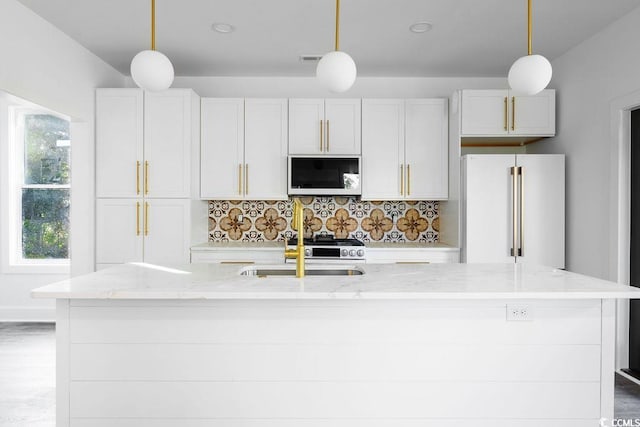 kitchen featuring light stone counters, white microwave, a center island with sink, freestanding refrigerator, and decorative backsplash