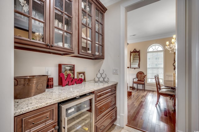 bar with light stone countertops, wine cooler, a chandelier, light wood-type flooring, and ornamental molding