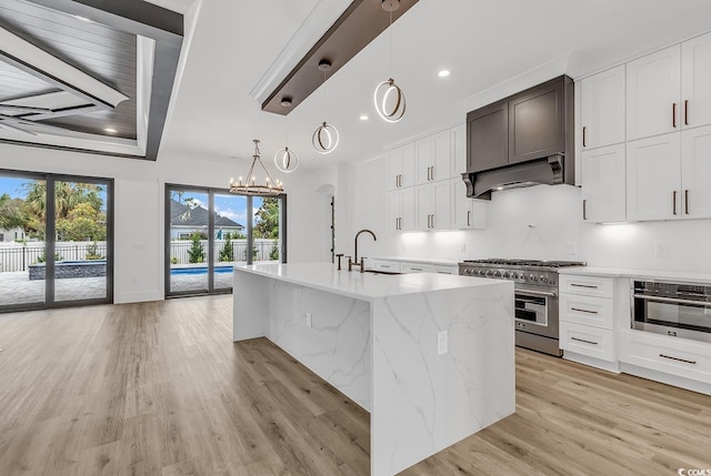 kitchen featuring stainless steel appliances, light wood finished floors, light stone counters, a center island with sink, and a chandelier