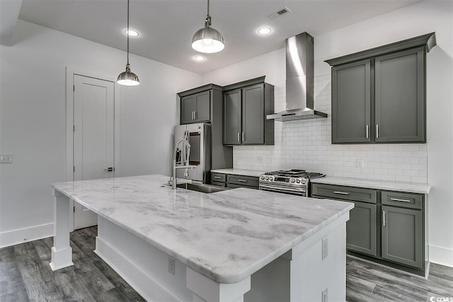 kitchen featuring wall chimney range hood, a center island with sink, hanging light fixtures, appliances with stainless steel finishes, and dark wood-type flooring