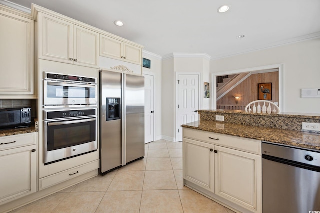 kitchen with dark stone counters, appliances with stainless steel finishes, crown molding, cream cabinetry, and light tile patterned flooring