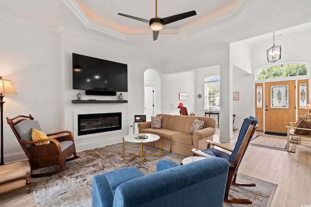 living room with light wood-type flooring, ceiling fan with notable chandelier, a tray ceiling, and crown molding