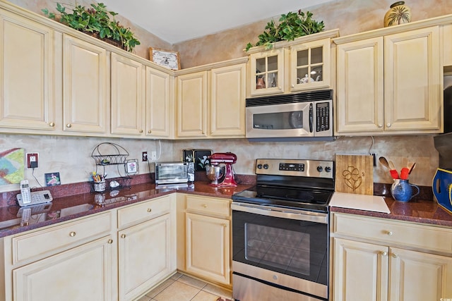 kitchen featuring backsplash, dark stone countertops, cream cabinetry, light tile patterned flooring, and appliances with stainless steel finishes