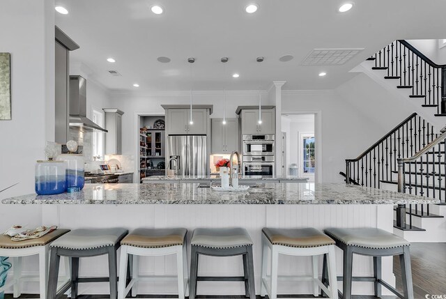 kitchen featuring pendant lighting, gray cabinetry, stainless steel appliances, a kitchen bar, and wall chimney exhaust hood