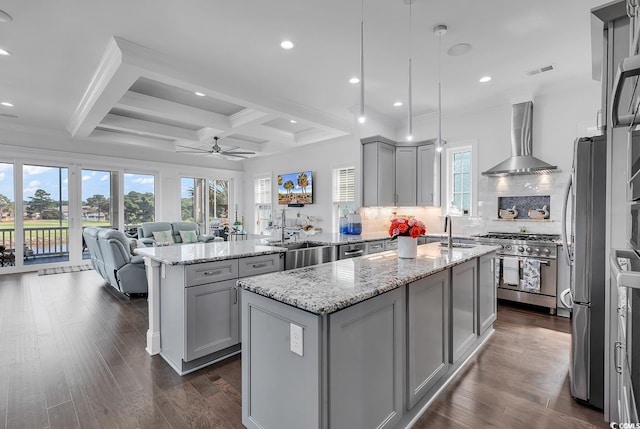 kitchen featuring wall chimney range hood, hanging light fixtures, stainless steel appliances, light stone countertops, and an island with sink